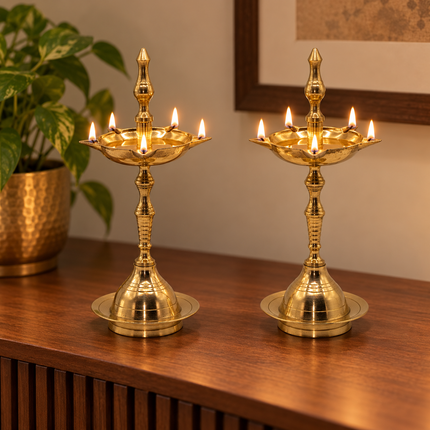 Two brass nilavilakku lamps with lit jyotis, displayed on a wooden table next to a potted plant in a warm indoor setting.