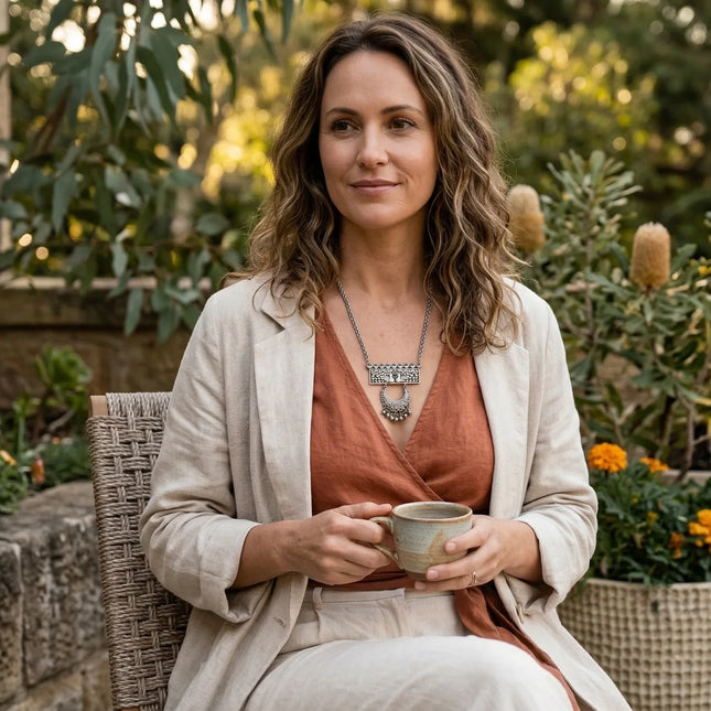 Woman sitting outdoors holding a cup, surrounded by plants and stone wall.