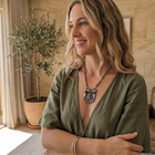 Woman in a green top standing in a room with a plant and a brown chair.
