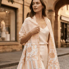 Woman walking on a street wearing a light pink embroidered shawl over a white outfit.