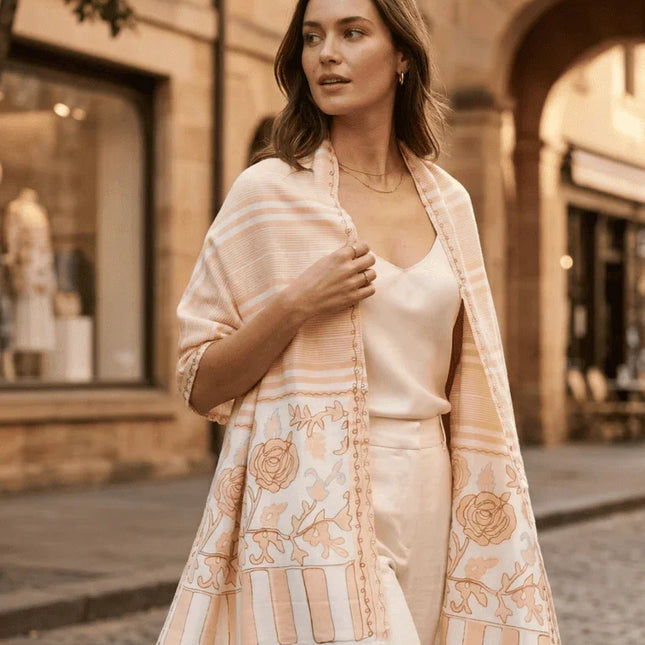 Woman walking on a street wearing a light pink embroidered shawl over a white outfit.